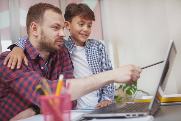 Young boy embracing his teacher while working on laptop together at school. Bearded mature man working at elementary school, explaining assignment to his young student, copy space - Photo, Image