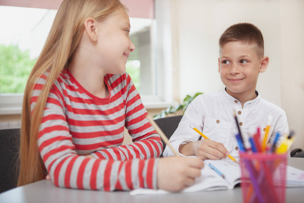 Прекрасні молоді однокласники посміхаються один одному під час малювання в класі. Веселий молодий хлопчик насолоджується малюванням у школі зі своїм другом
 - Фото, зображення