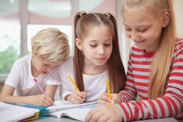 Three lovely shoolgirls chatting during elementary school drawing lesson. Beautiful little girl looking at her friend sketching. Education, friendship, interaction concept - Фото, зображення