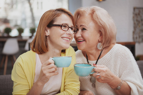 Cheerful senior mother whispering to her daughter. Happy elderly woman enjoying talking to her adult daughter over cup of coffee - Фото, зображення