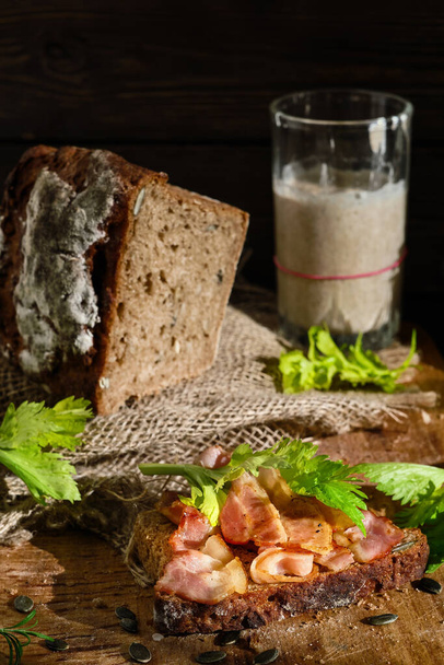 Sendvič se slaninou a celerovými listy na plátek žitného chleba z kvásku s dýňovými semínky. Zdravé jídlo koncept, tradiční řemeslný chléb, sklenice s kysaným těstem na stole. - Fotografie, Obrázek