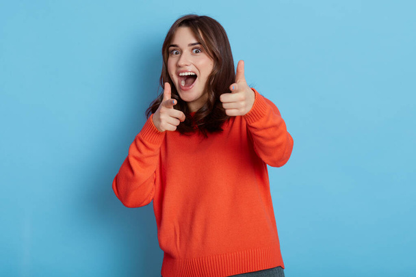 Good looking attractive brunette girl, look camera with smiling expression, pointing to camera with fore finger, introduce amazing product, being excited, keeps mouth opened, stands against blue wall. - Valokuva, kuva