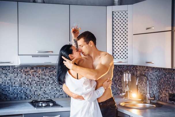 Slavíme Valentýna. Milující heterosexuální pár s krásnými nahými postavami a těly objetí, polibek, v kuchyni. Dívka drží sklenici vína v rukou, užívá si vášnivého okamžiku. - Fotografie, Obrázek