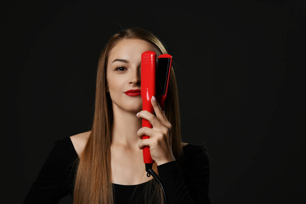 Young smiling beautiful woman with long silky straight hair in black clothes holding red hair straightener in hand over one eye - Fotó, kép