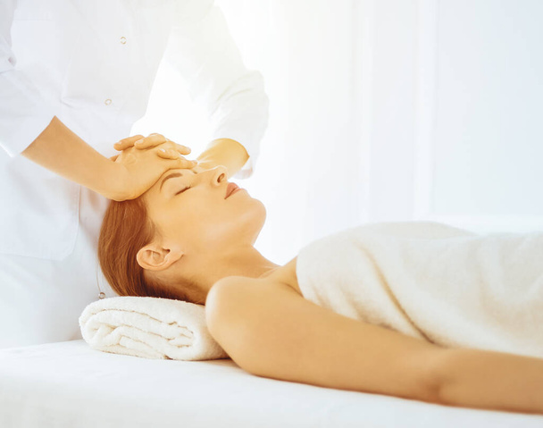 Beautiful woman enjoying facial massage with closed eyes in sunny spa center. Relaxing treatment concept in medicine - Фото, зображення