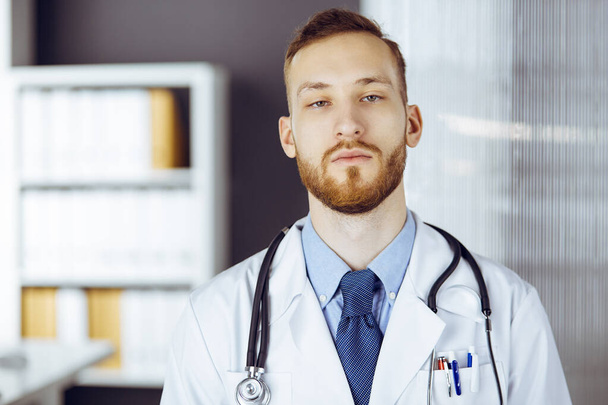 Red-bearded doctor standing straight in clinic near his working place. Portrait of physician. Medicine and healthcare concept - Foto, immagini