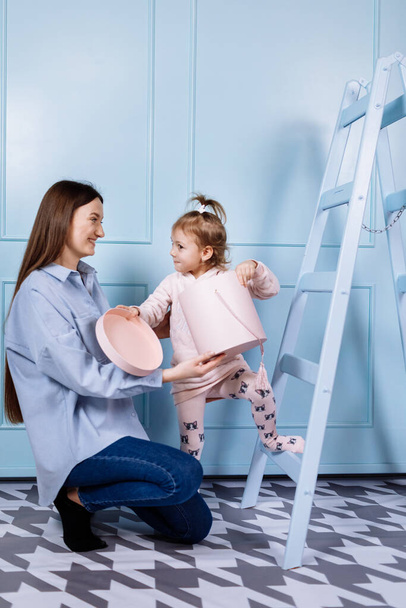 Feliz familia amorosa. joven madre en azul y su hija pequeña en ropa rosa jugando con caja de regalo rosa y escalera azul sobre fondo de pared azul. madres felices, día del bebé - Foto, Imagen