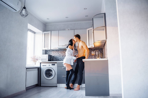 Attractive, passionate, handsome girl and guy having love in the kitchen. Boyfriend with naked muscular torso body gently hugs woman in a white shirt, pressing her against the wall. - Fotoğraf, Görsel