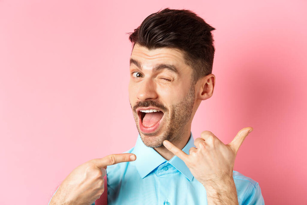 Cheeky and funny guy showing phone gesture, asking to call him and winking, telling dial his number, standing over pink background - Photo, image
