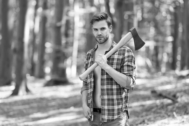 Unshaven handsome man in open plaid shirt with jeans carry large splitting axe in summer forest natural landscape, lumberman - Photo, image