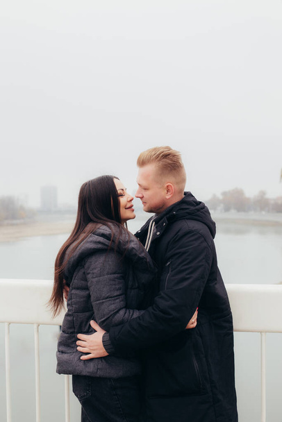 A young couple in warm clothes hugs and looks at each other against the background of a foggy landscape. High quality photo - Foto, imagen