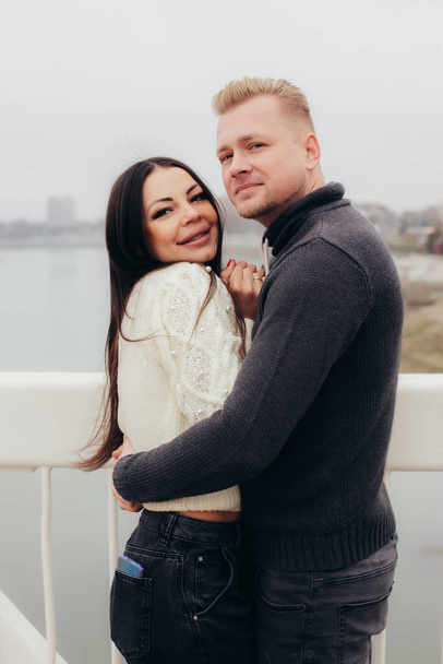 In love couple in sweaters cuddles smiling on the bridge, in the background of the river and high houses. High quality photo - Foto, afbeelding