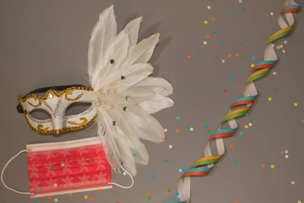 Bílá karnevalová maska s třpytkami, červená chirurgická maska, barevné konfety a fáborky na šedém pozadí, v popředí. - Fotografie, Obrázek