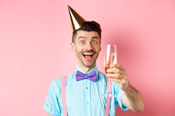 Holidays and celebration concept. Excited birthday guy raising glass of champagne, making toast or speech, standing in party hat and having fun on pink background - Fotoğraf, Görsel