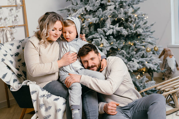 Christmas Family Happiness Portrait of dad, pregnant mom and little son  sitting armchair at home near Christmas tree hug smile European young adult family holiday morning - Foto, Imagen