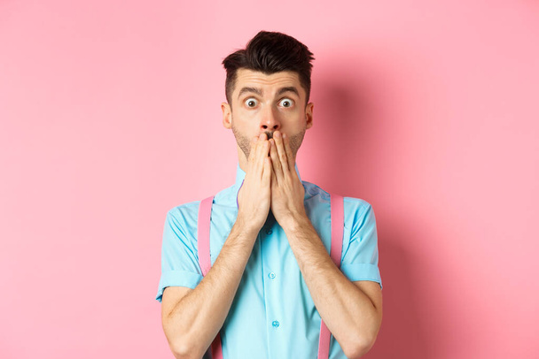 Σοκαρισμένος άντρας να κοιτάζει τρομαγμένος, να καλύπτει το στόμα με τα χέρια και να κοιτάζει την κάμερα, να στέκεται σε ροζ φόντο - Φωτογραφία, εικόνα