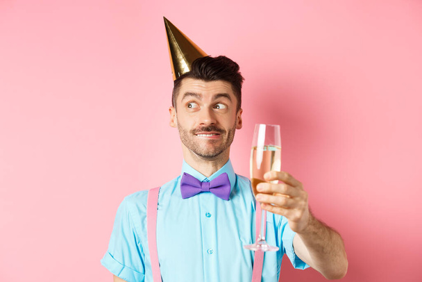 Ünnepek és ünnepség koncepció. Vidám fiatalember ünnepli születésnapját party kalap, gondolkodás beszéd, emelés pohár chamapgne pirítós, rózsaszín háttér - Fotó, kép