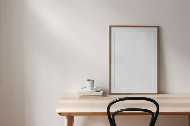 Breakfast still life scene. Cup of coffee, books and empty picture frame mockup on wooden desk, table. Elegant working space, home office concept. Scandinavian interior design. Room in sunlight. - 写真・画像