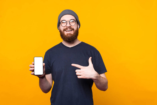 Neşeli sakallı hippi adam sarı arka planda akıllı telefon ekranını işaret ediyor. - Fotoğraf, Görsel