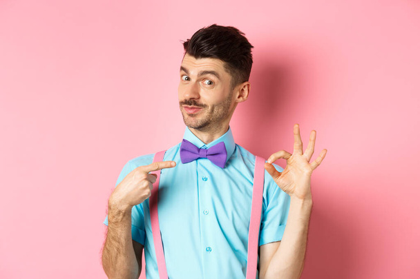 Image of young handsome guy in bow-tie and holiday outfit, pointing at himself and showing okay gesture, self-promoting on pink background - Фото, изображение