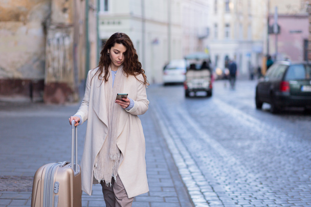 Beautiful brunette woman walking in a new city with a phone in her hands and a suitcase, a tourist discovers new cities - Foto, imagen