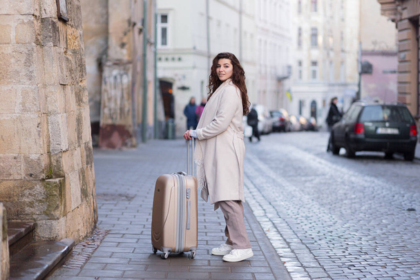 Όμορφη μελαχρινή γυναίκα περπατά σε μια νέα πόλη με ένα τηλέφωνο στα χέρια της και μια βαλίτσα, ένας τουρίστας ανακαλύπτει νέες πόλεις - Φωτογραφία, εικόνα