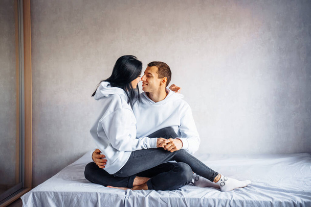 Beautiful young couple in the same white hoodie and black pants lie on the bed hugging. Dreaming of happy future together. Enjoying each other's company. Man gently hugs his beloved from back. - Photo, Image