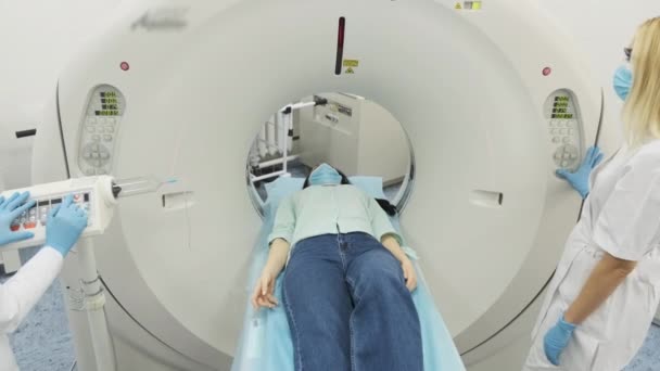 Paciente femenina sometida a tomografía computarizada o resonancia magnética bajo la supervisión de dos radiólogos calificados en máscara y guantes en la clínica médica moderna. Paciente acostado en una mesa de tomografía computarizada o resonancia magnética - Metraje, vídeo