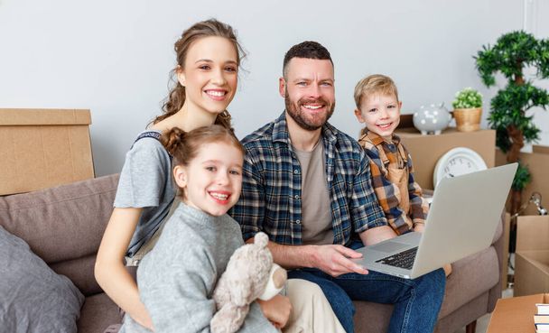onnellinen perheen äiti isä ja lapset muuttavat uuteen asuntoon ja istuvat sohvalla laatikoiden joukossa, jotka opiskelevat tietoa Internetissä kannettavalla tietokoneella - Valokuva, kuva