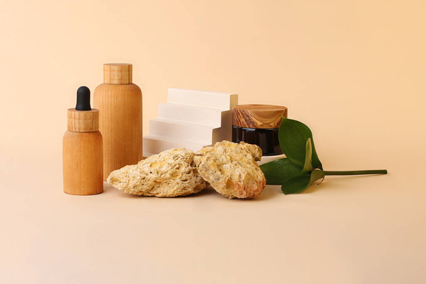 Eco friendly bottles from the natural wood and brown glass near the geometrical pedestal.Fresh natural leafs and decorative stones on the background.Concept of the organic,zero waste cosmetics. - Photo, image