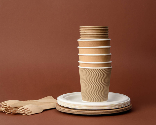 taza de papel, platos blancos y tenedores y cuchillos de madera sobre un fondo marrón. Concepto de rechazo de plástico, cero residuos  - Foto, imagen