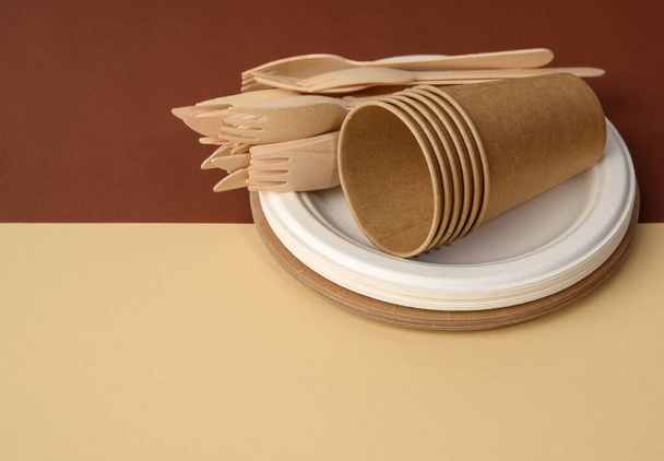 paper cup and wooden fork, empty round brown disposable plate made from recycled materials on a brown background. Concept of the absence of non-recyclable garbage, rejection of plastic - Photo, Image