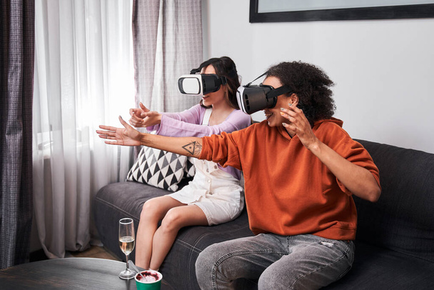Las mejores amigas sentadas en un sofá acogedor y usando gafas de realidad virtual - Foto, Imagen