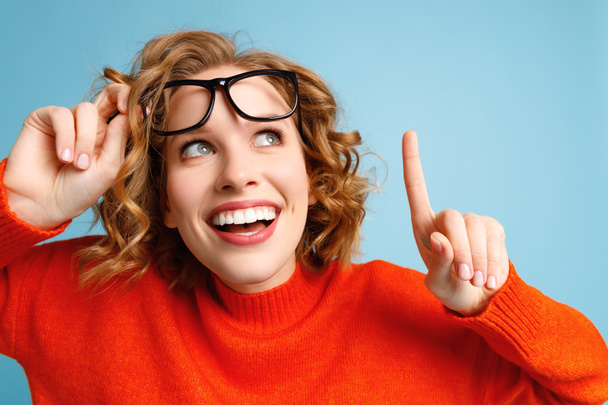 Feliz joven hembra levantando gafas con estilo y sonriendo mientras señala hacia arriba contra fondo azul - Foto, Imagen