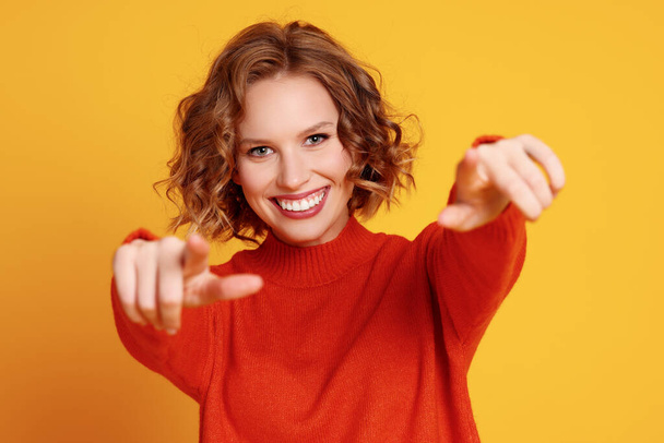 Счастливая молодая женщина в стильном красном свитере улыбается и указывает на камеру на желтом фоне - Фото, изображение