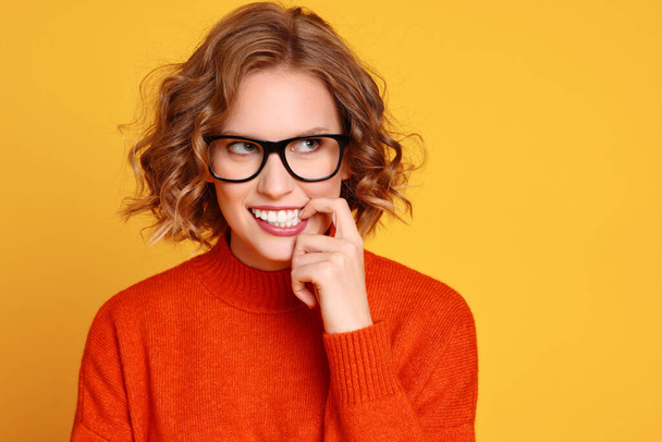 Young female in trendy red sweater and glasses biting finger and looking away while dreaming against yellow background - Photo, Image