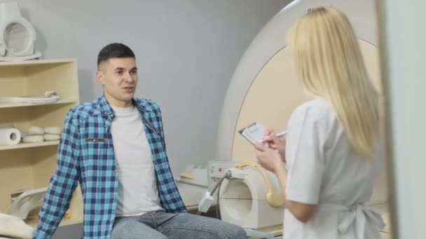 Жінка-лікар радіолог має діалог з молодим чоловіком-пацієнтом, питає і відзначає медичну історію пацієнта, скарги в сучасній клініці поряд з сучасним МРТ закритого типу. - Кадри, відео