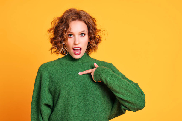Überraschte junge Frau im trendigen grünen Pullover blickt mit geöffnetem Mund in die Kamera und zeigt auf die Brust vor gelbem Hintergrund - Foto, Bild