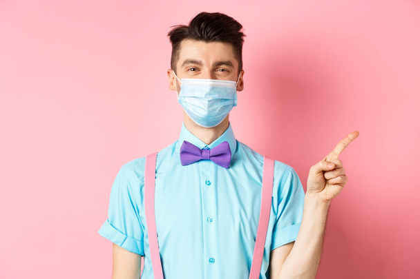 Covid-19, pandemian ja terveyden käsite. Hymyilevä valkoihoinen mies lääketieteellisessä naamiossa osoittaa sormella oikealle, näyttää mainosta, seisoo vaaleanpunaisella taustalla - Valokuva, kuva