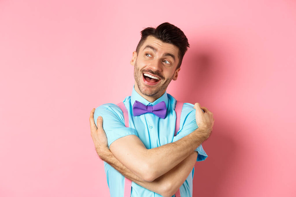 Счастливый и позитивный парень в галстуке-бабочке, обнимающий себя и выглядящий слева с веселой улыбкой, стоящий на розовом фоне - Фото, изображение