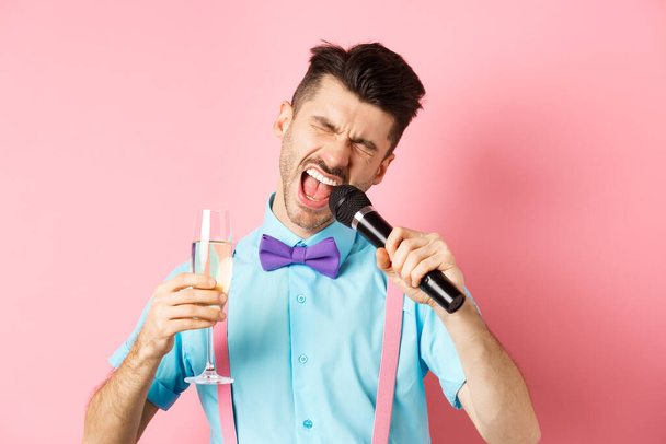 Party and festive events concept. Drunk funny guy singing in microphone and drinking champagne from glass, having fun at karaoke bar, standing on pink background - Fotó, kép