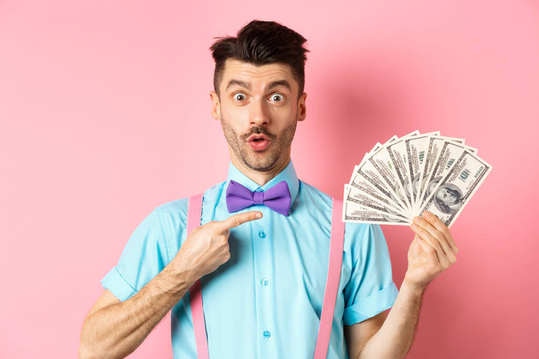 Niesamowity człowiek pokazujący wielką nagrodę pieniężną, wskazujący na dolary i mówiący wow, patrzący pod wrażeniem kamery, stojący nad różowym tłem - Zdjęcie, obraz