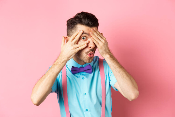 Заинтригованный смешной парень в галстуке-бабочке, подглядывающий сквозь пальцы на что-то интересное, проверяющий что-то классное, стоящий на розовом фоне - Фото, изображение