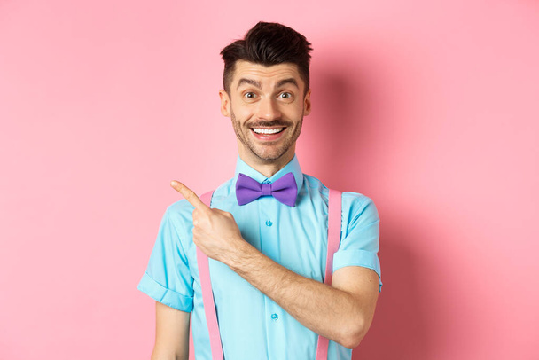 Portrait of handsome happy man showing logo, pointing left and smiling at camera, recommending shop offer, standing on pink background - Foto, Imagem