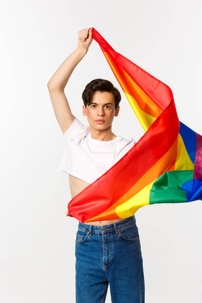 Vertical view of happy queer person in crop top and jeans waving raised rainbow flag, celebrating lgbtq holiday, standing over white background - Fotó, kép