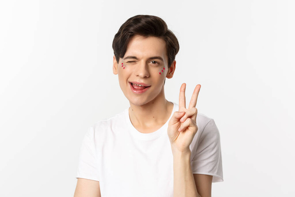 Mensen, lgbtq gemeenschap en lifestyle concept. gelukkig en schattig gay man met glitter op gezicht tonen vrede teken en glimlachen vieren trots vakantie wit achtergrond - Foto, afbeelding