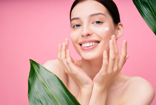 Smiling tender cute girl with face cream on her cheek posing on a pink studio background, near the face of green leaves. Cosmetic product for facial beauty and natural skin care. - 写真・画像