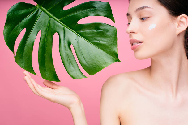 Close-up, portrait of a tender young caucasian girl with a nourishing face cream on her cheek, touching a green monstera leaf near her face, nude. Natural beauty and organic cosmetics concept. - Foto, Imagem