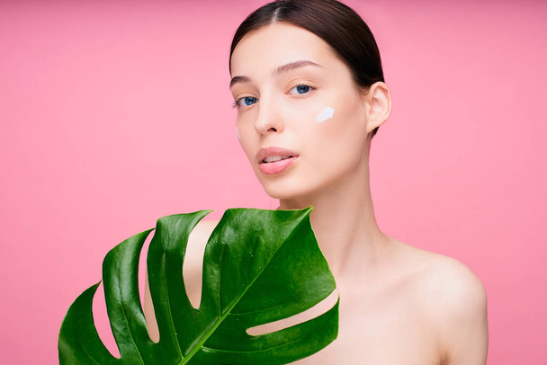 Cute lovely tender young Caucasian girl with a nourishing face cream on her cheek, looks straight, covers part of the frame with a large green leaf, has dark hair, isolated on a pink background. - Φωτογραφία, εικόνα