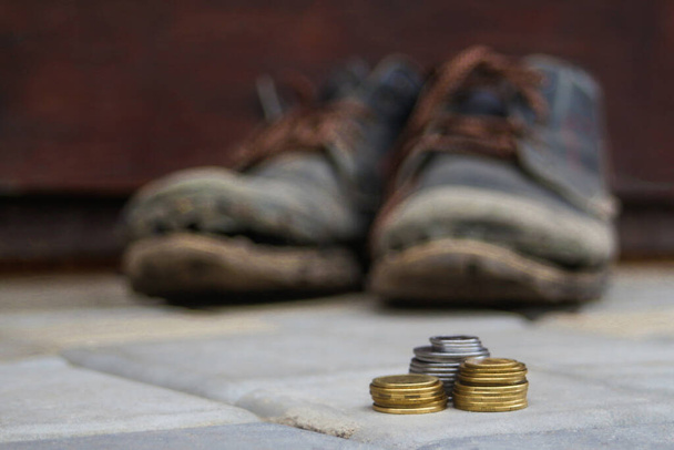 Ułożone monety na rozmytym tle pary starych brudnych butów. Koncepcja ubóstwa, bezdomności, braku pieniędzy. Szare kostki brukowe. Skupienie selektywne - Zdjęcie, obraz